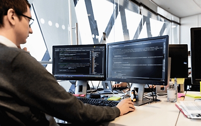 Een persoon die achter een bureau werkt met twee desktopmonitors waarop code wordt weergegeven