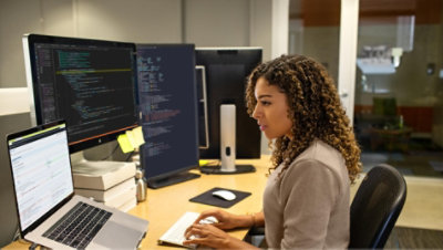 Eine Person, die am Schreibtisch mit einem Laptop und mehreren Monitoren arbeitet