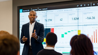 A person giving a presentation in front of a small crowd with Dynamics 365 being displayed on the wall behind them