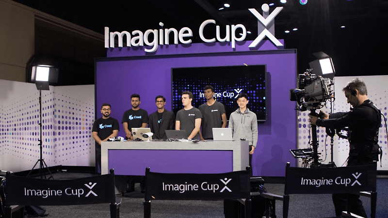Enam orang berpartisipasi dalam Imagine Cup sedang berdiri di meja dan difilmkan.