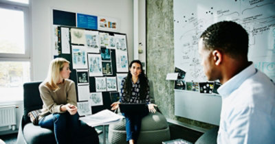 Troje kolega razgovaraju u moderno opremljenoj kancelariji, a na zidu iza njih se nalazi bela tabla sa dijagramima i zakačeni nacrti