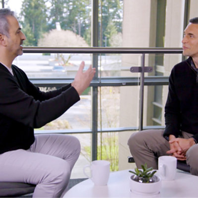 Two men sit across from each other at a small table with coffee cups and a potted plant, engaged in conversation 