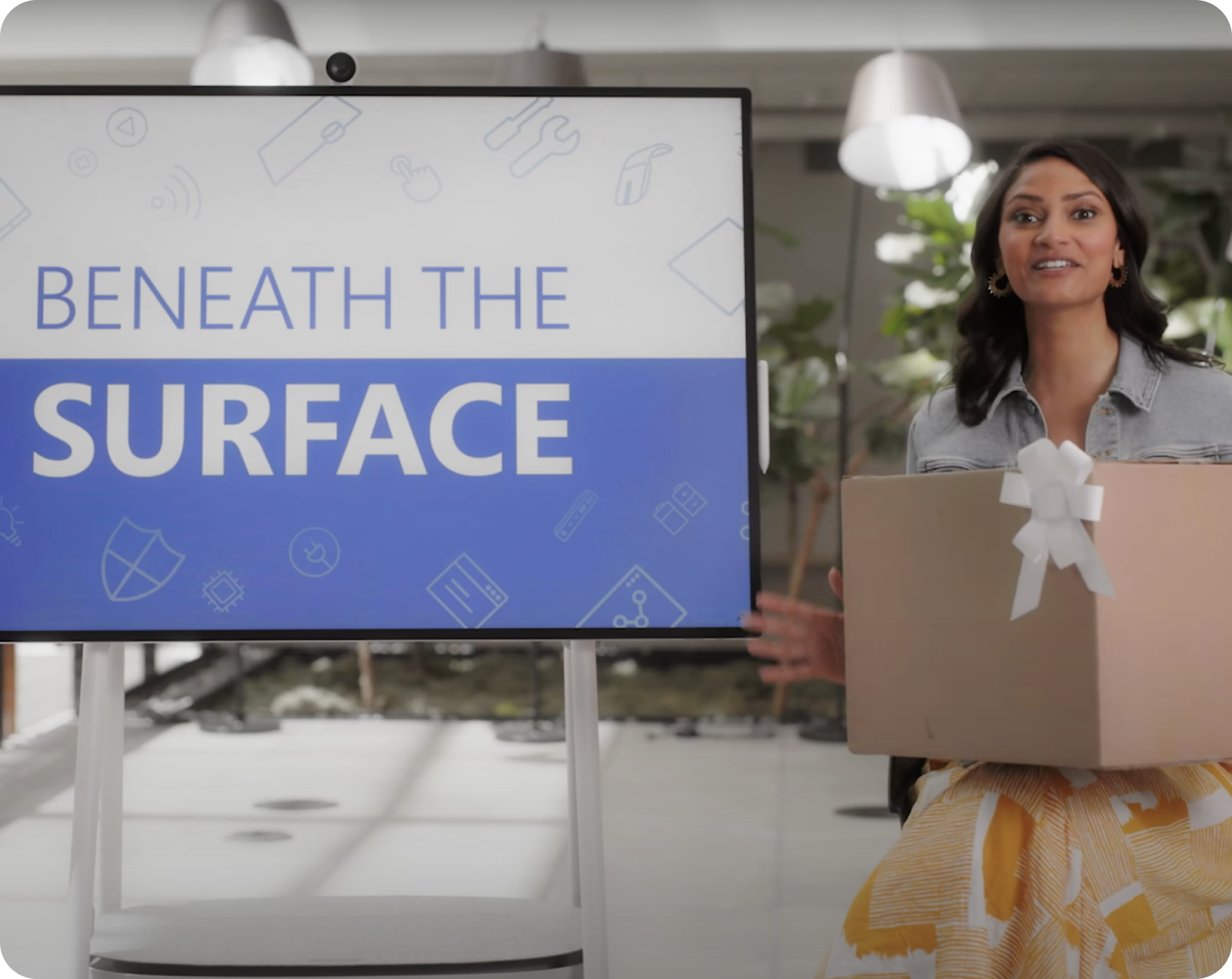 A woman in a dress stands next to a screen displaying "Beneath the Surface" holding a wrapped box.