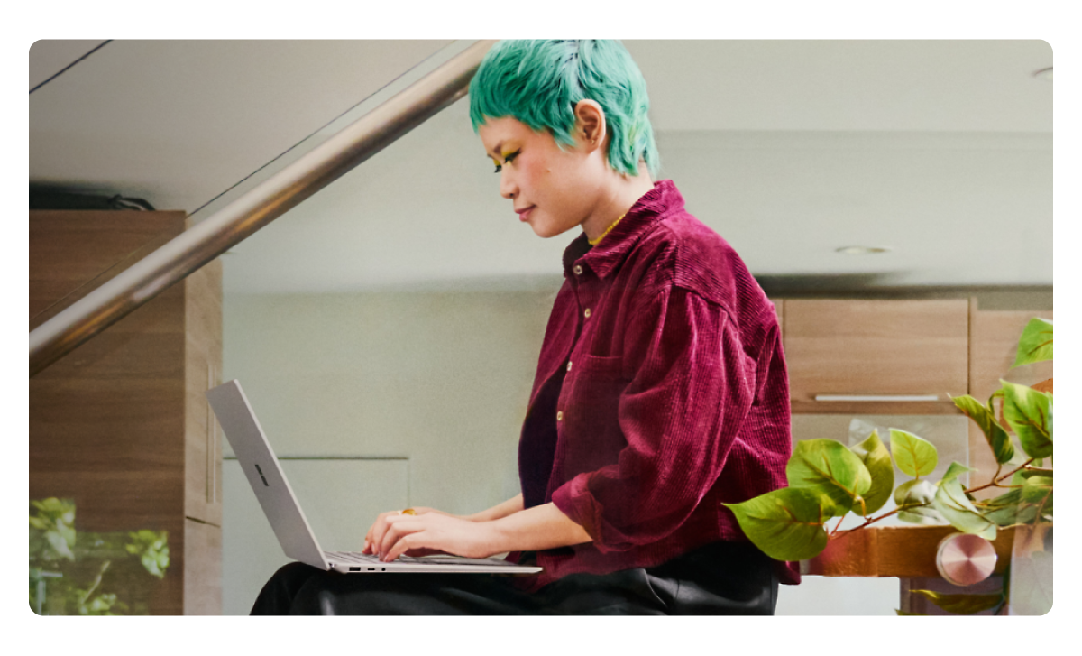 A person with short blue hair types on a laptop while seated indoors. They are wearing a red shirt and have green makeup.