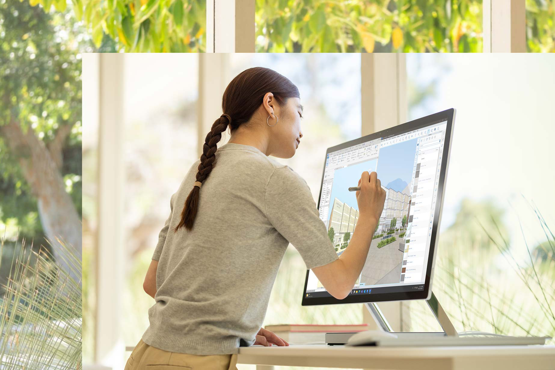 街並みの一部が見える窓の前で Surface Studio 2+ に向かって座る男性。