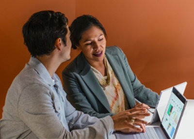 Two people are sitting at a table with laptops open, discussing work in an office setting