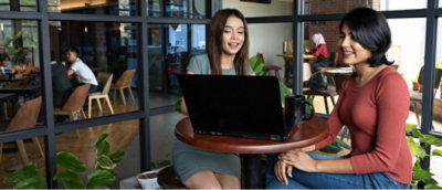 Two individuals sitting at a round table with a laptop in a busy cafe setting.