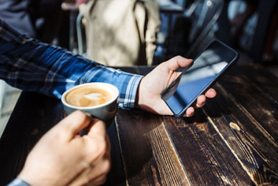 Osoba u plavoj kariranoj košulji sedi za drvenim stolom i u jednoj ruci drži šolju kafe, a u drugoj pametni telefon.