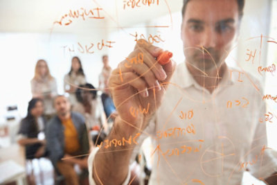 Muškarac u beloj košulji piše narandžastim markerom na providnoj tabli, a grupa ljudi ga posmatra otpozadi