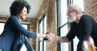 Muškarac i žena se rukuju u jarko osvetljenoj prostoriji sa zidovima od cigala i velikim prozorima. Oboje se osmehuju.