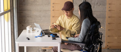 A woman in a wheelchair talking to a man at a table with a laptop.