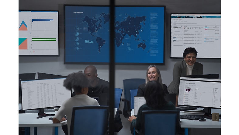 Personas hablando con varios escritorios abiertos y una tabla de gráficos abierta, mapa del mundo en un panel de pantalla grande