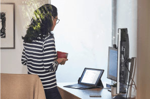 A person standing, holding a mug and looking at a tablet in laptop mode on a desk