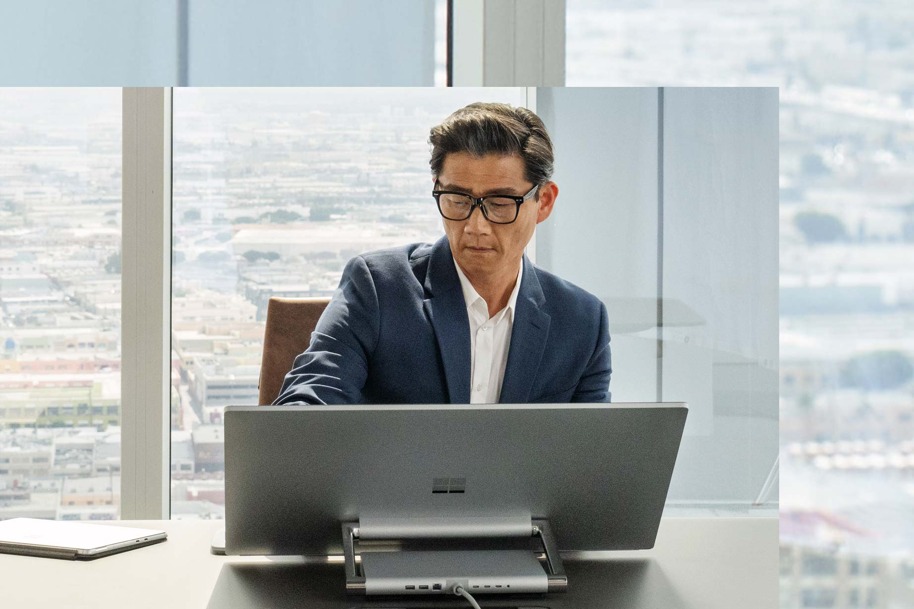 Un homme assis devant son Surface Studio 2+ face à une fenêtre derrière laquelle se profile la ville.