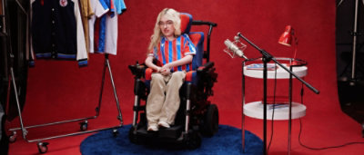 Person using a powered wheelchair in a studio setting with a microphone, table, and sports jerseys in the background.