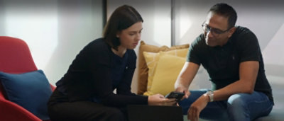 A man and woman are seated and engaged in a discussion while looking at laptops.