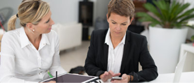 Two professionals in business attire sitting at a table with one holding a pen and paper and the other using a smartphone.