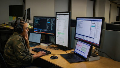 Eine Person, die Kopfhörer trägt und an ihrem Schreibtisch an einem Laptop arbeitet, der an mehrere Desktopmonitore angeschlossen ist