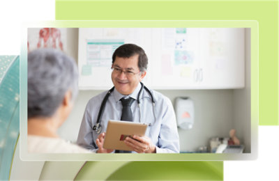A doctor in a white coat sits at a desk, speaking on a phone. A computer, books, and a lamp are on the desk.