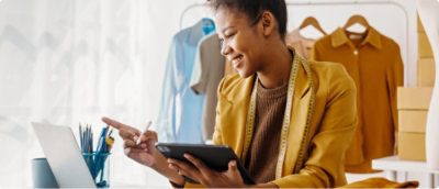 Une personne en veste jaune utilise une tablette avec un stylet à un bureau avec un ordinateur portable et des fournitures de bureau, avec des étagères de vêtements à l’arrière-plan.