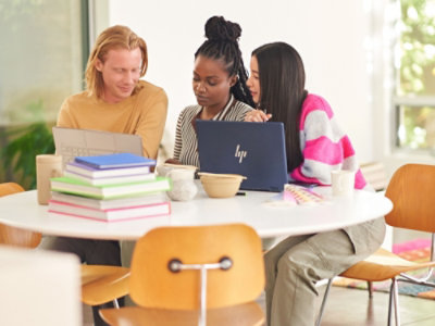 3 人がノート PCと本を備えたテーブルに座り、明るい部屋で一緒に作業をしています。
