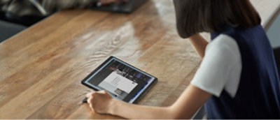 Person, die mit einem Stift auf einem Tablet an einem Holztisch arbeitet.