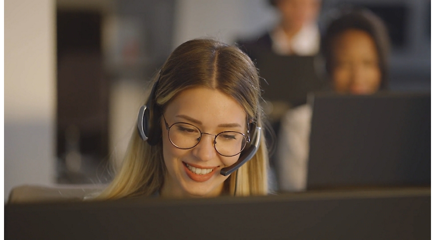 Una mujer sentada en la oficina delante de su escritorio y hablando mientras lleva auriculares