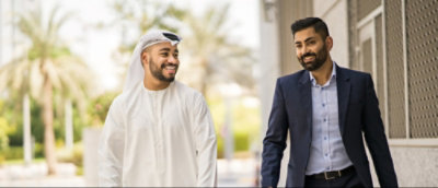 Two men walking side by side, one in traditional Emirati attire and the other in a business suit, in an outdoor setting with palm trees in the background.