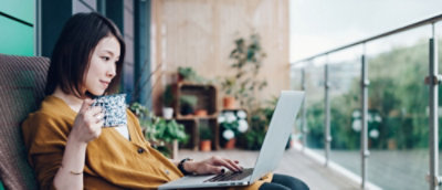 Person in a yellow top sitting on a couch working on a laptop in a modern room with large windows.