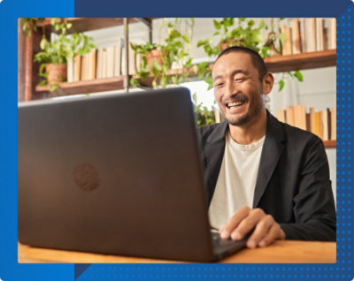 Eine lächelnde Person, die an einem Tisch mit einem Laptop arbeitet. Im Hintergrund sind Bücherregale und Topfpflanzen zu sehen.