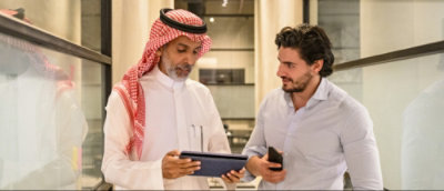 Two men, one in traditional Arab attire and the other in a casual shirt, standing and discussing over a tablet.