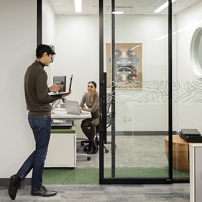 Muž vstupující do kanceláře držící přenosný počítač a ženy uvnitř ho pozdraví.