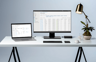 Laptop and desktop are placed on a table with some reports open and a table lamp next to it.