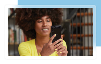 A person with curly hair uses a smartphone, wearing a yellow top and nail polish, in a room with bookshelves.
