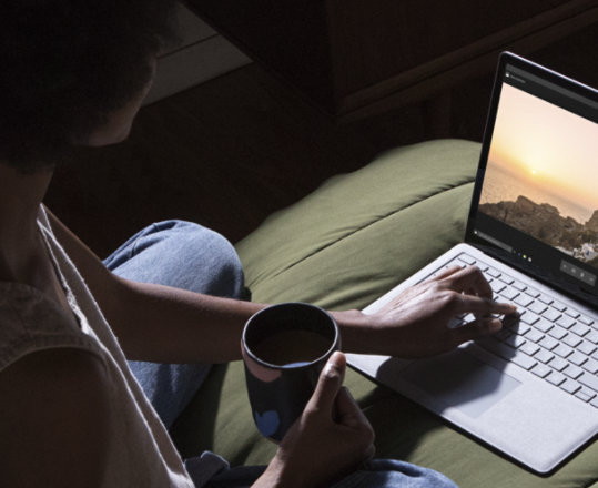 Photograph of person sitting cross-legged on a bed drinking something from a mug and using a laptop