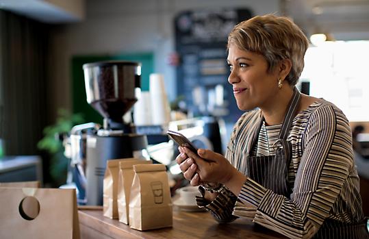A person working at a coffee shop leaning against the counter and smiling while holding a mobile device.