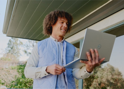 A person holding and looking at a laptop outside, standing under a roof with trees in the background.