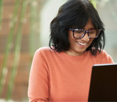En kvinde med sort hår og briller, kigger på en bærbar computer.