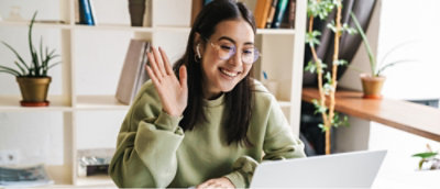 緑のセーターを着た、眼鏡をかけた人がノート PCの前のデスクに座り、笑顔で手を振っている。