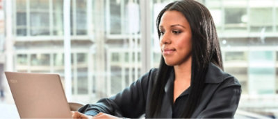 Person in a black blazer working on a laptop with a windowed office building in the background.