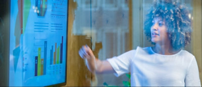 A person in a white shirt gestures towards a screen displaying various bar graphs and data visualizations.